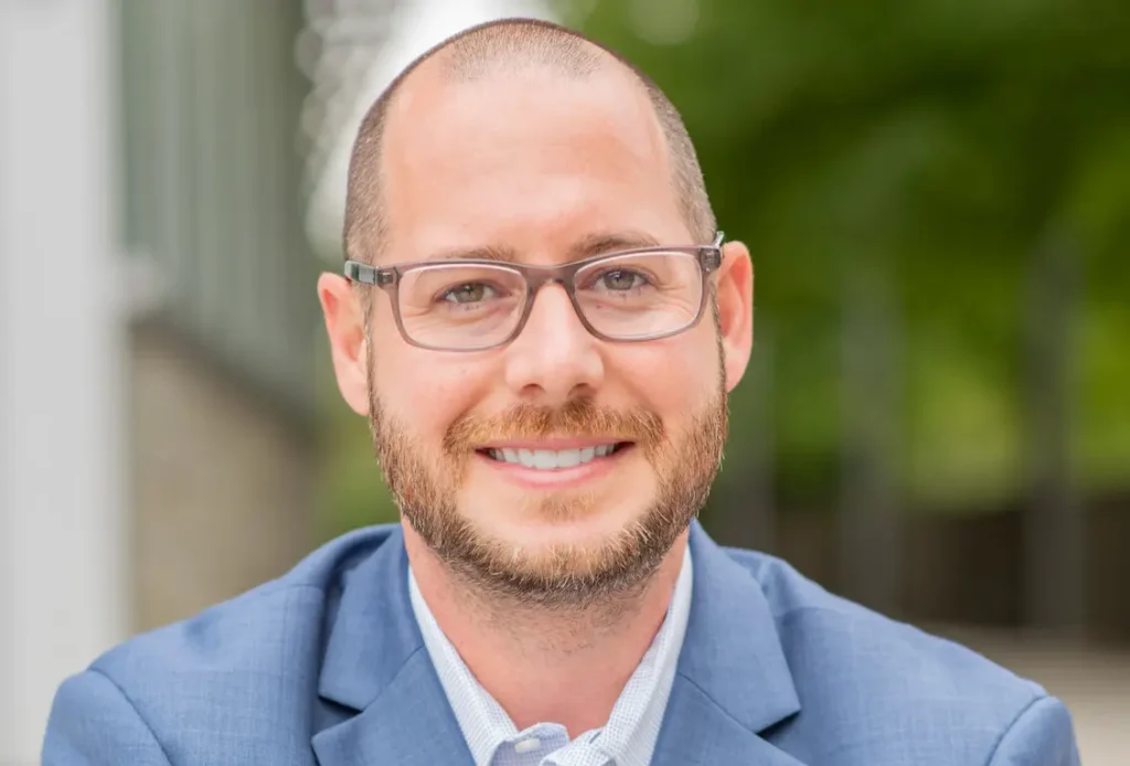 A person with glasses and a beard smiles in an outdoor setting, wearing a light blue shirt and a blue blazer.